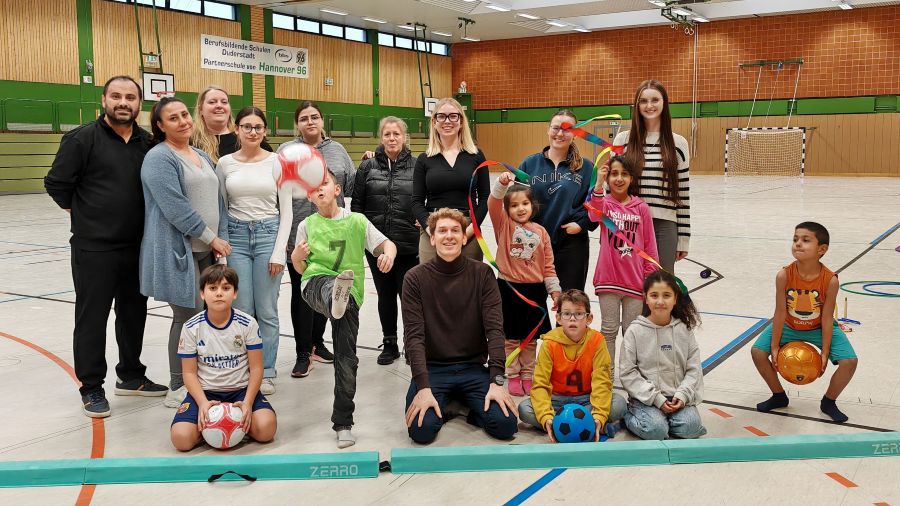 Ein Gruppe Erwachsener und Kinder stehen in einer Sporthalle, einige Kinder halten einen Ball, ein Jung schießt einen Ball in die Luft.