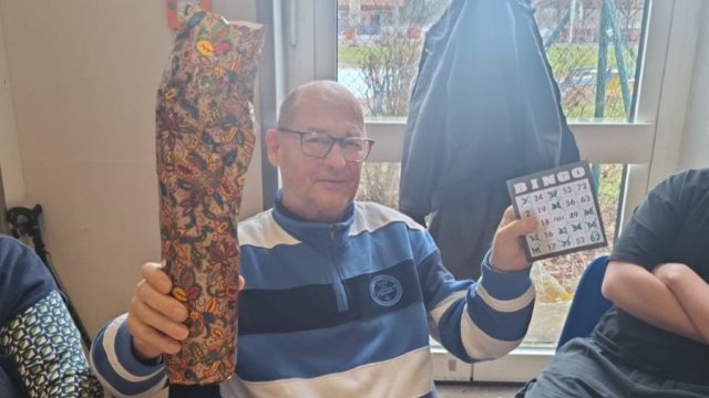 Ein Mann hält in der rechten Hand ein eingepacktes Geschenk, in der linken Hand eine Bingokarte.